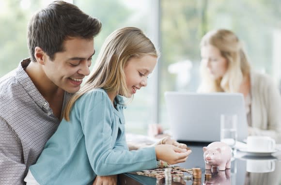 A smiling man watches a girl with a piggy bank and stacks of coins.