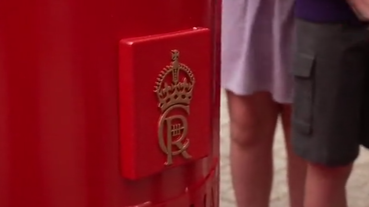 A close up of King Charles' cypher, which has been applied in gold onto the red post box, with school pupils standing in the background