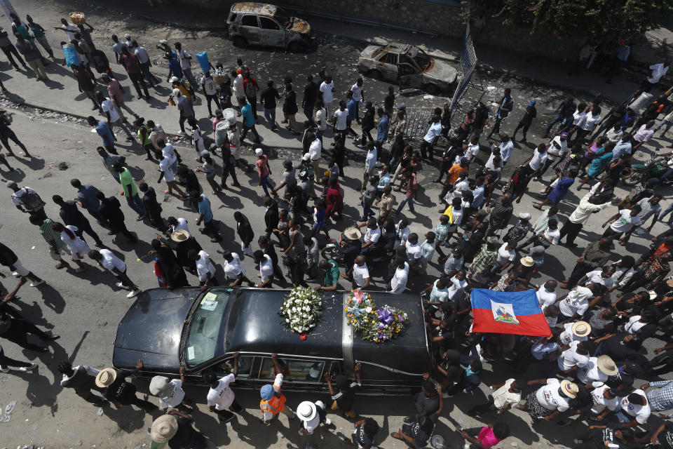 The funeral procession accompanying the body of murdered community leader Josemano "Badou" Victorieux to the cemetery passes by cars burnt during recent demonstrations in the Delmas area of Port-au-Prince, Haiti, Wednesday, Oct. 16, 2019. Funerals for 11 of at least 20 people killed were held in six cities, including the capital of Port-au-Prince, where at least two people were injured in a protest that broke out when presidential guards tried to block a road near where hundreds had gathered around the coffins of two victims. (AP Photo/Rebecca Blackwell)
