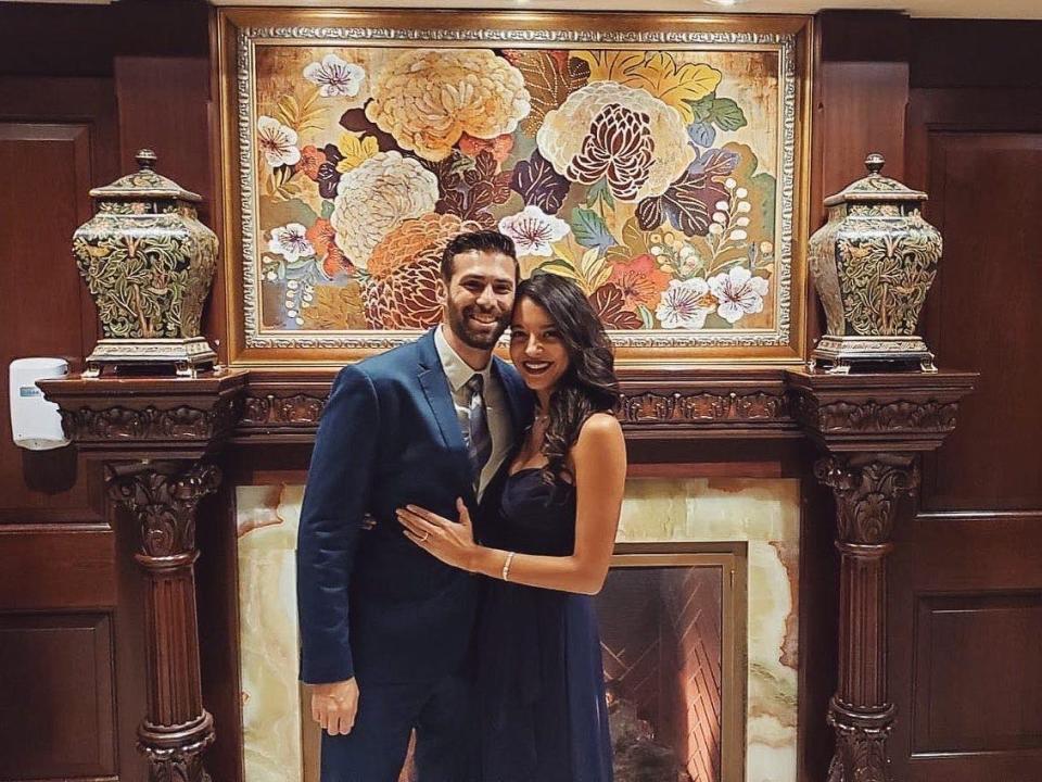 Joanna and her husband in formal attire, posing in front of an ornate fireplace.