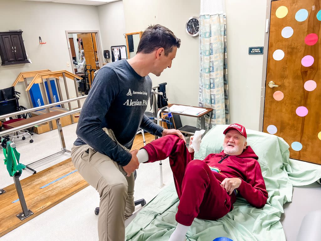 An elderly man in a track suit having his legs moved by a home care worker