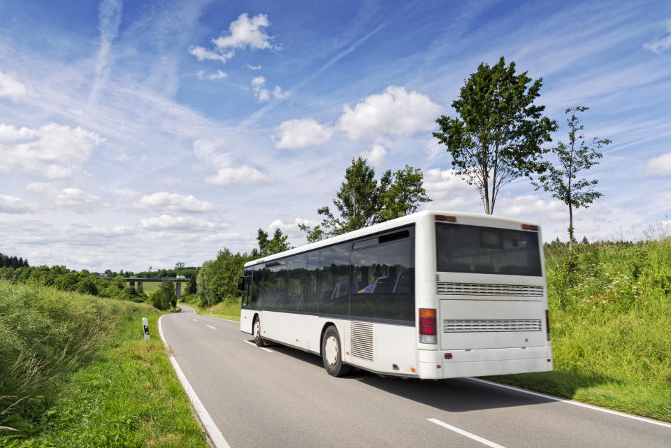 A white bus is driving on a rural road surrounded by green fields and trees under a cloudy sky