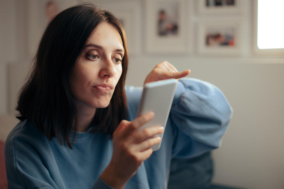 A woman in a blue sweatshirt holds a smartphone and points at the screen with her other hand