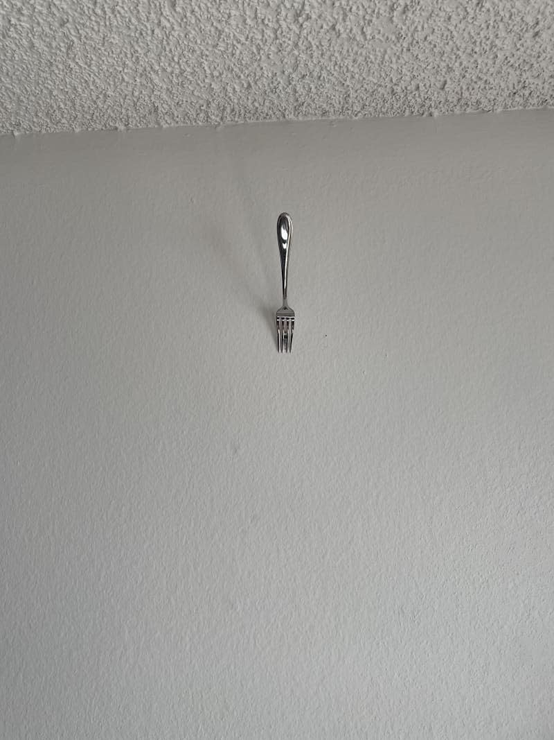 Fork slid over a nail that's been hammered into white-painted drywall, so that the nail is between two tines of the fork and the handle end is facing up.