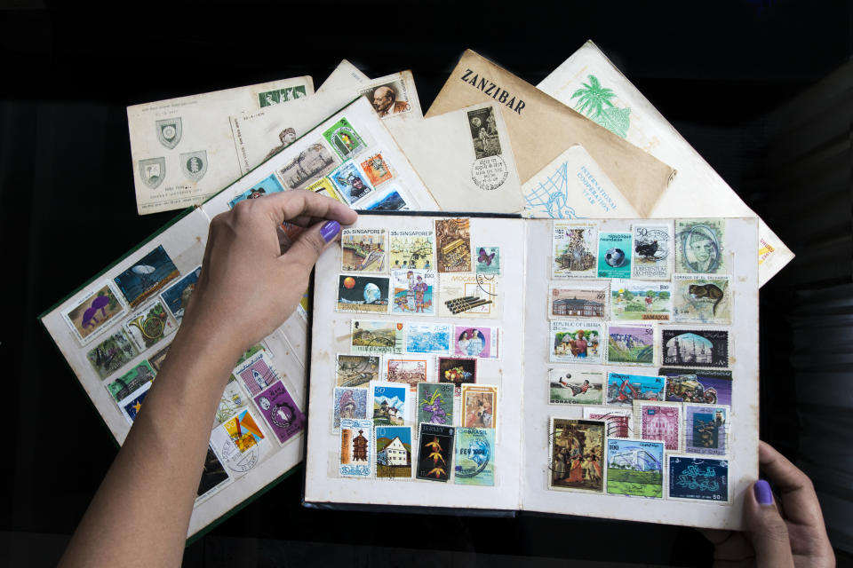 Hands are shown flipping through pages of a stamp collection album, with various colorful stamps laid out on a table alongside envelopes, one labeled "Zanzibar"