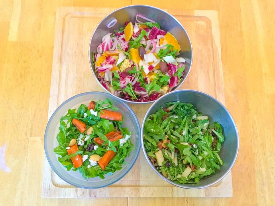 Bird's-eye-view of three salads on a wooden cutting board. The salads contain ingredients including arugula, carrots, orange wedges, purple cabbage, and apples.