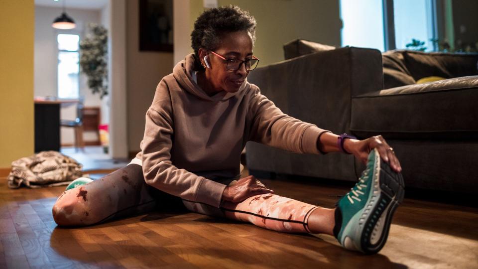 older black woman wearing a pink long sleeve workout set and sneakers stretches on the floor in her home while wearing in-ear headphones