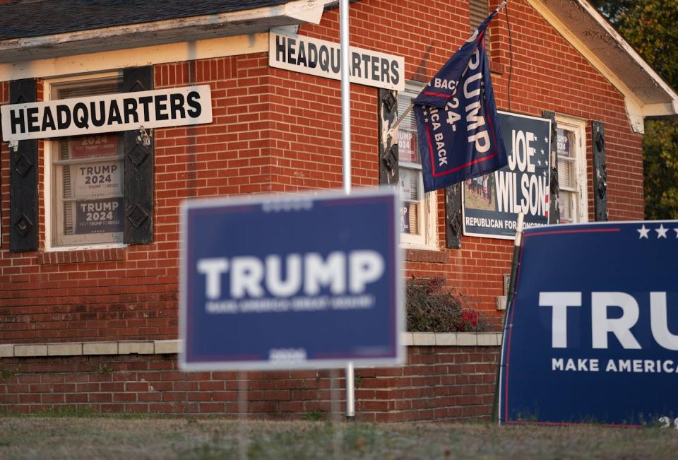 A series of blue lawn placards that say 'Trump' in white writing stand outside of a small brick building, which also has a Trump flag.