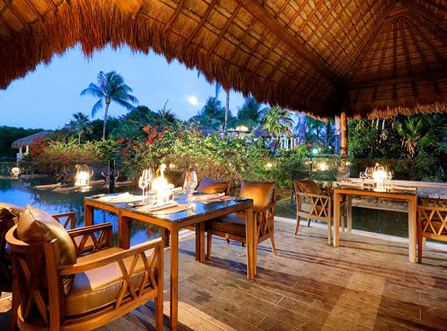 A photo of a dining table table for two by the lake surrounded by flowers and palm trees