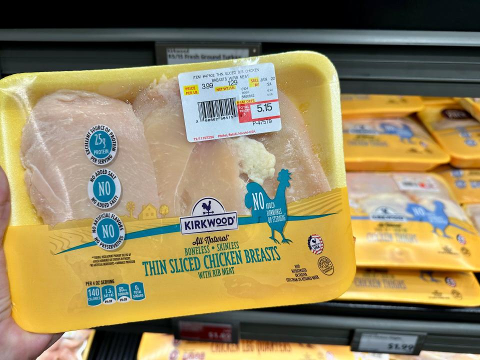 A hand holds a pack of chicken breasts with a white Kirkwood label and a $5.15 price tag