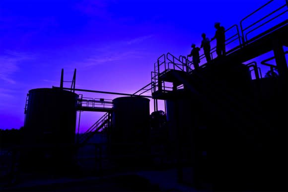 Oil storage facility at dusk with three workers looking from a railing overseeing the facility.