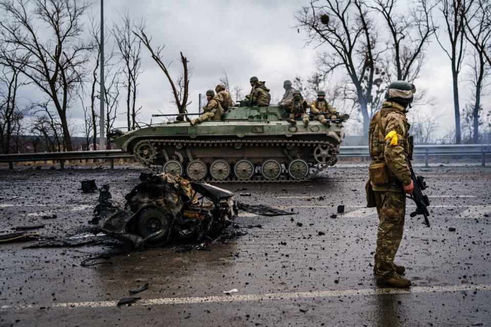 A military vehicle rolls past wreckage on a road.
