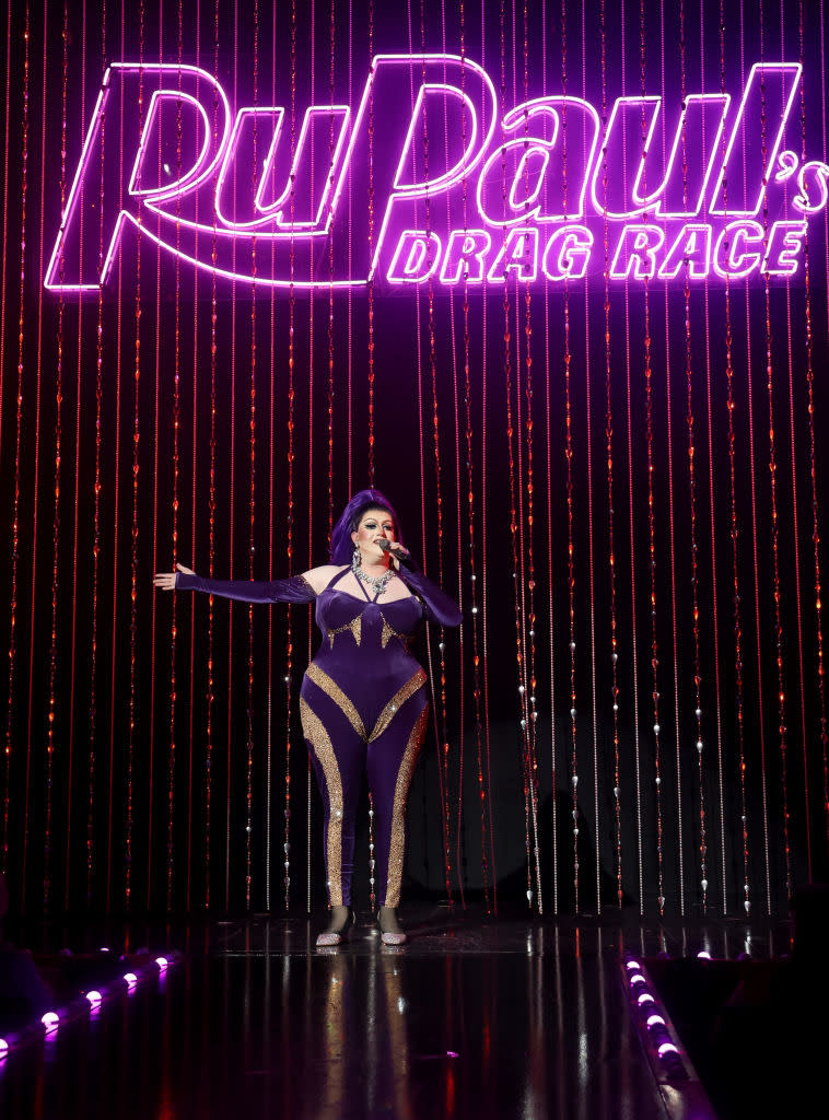 RuPaul stands on stage in a glamorous, form-fitting ensemble, performing under the "RuPaul's Drag Race" sign with sparkling backdrop