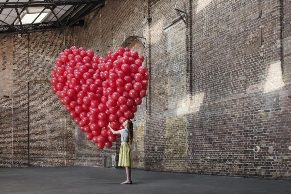 woman hugging a giant heart made of red helium balloons, standing inside an old brick building