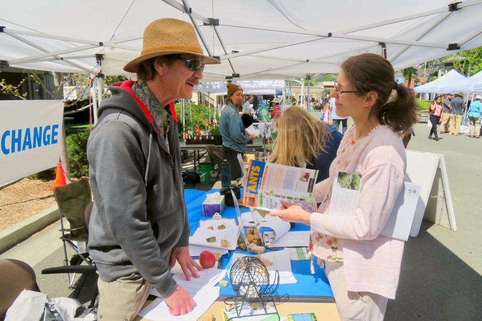 A man at an outdoor booth in what looks like a farmers market talks with a woman.