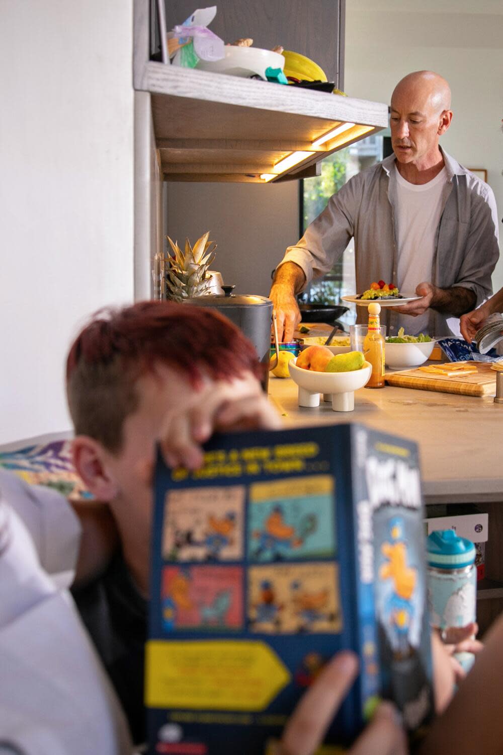 A man fixes dinner in the kitchen as a kid read in a nearby nook.