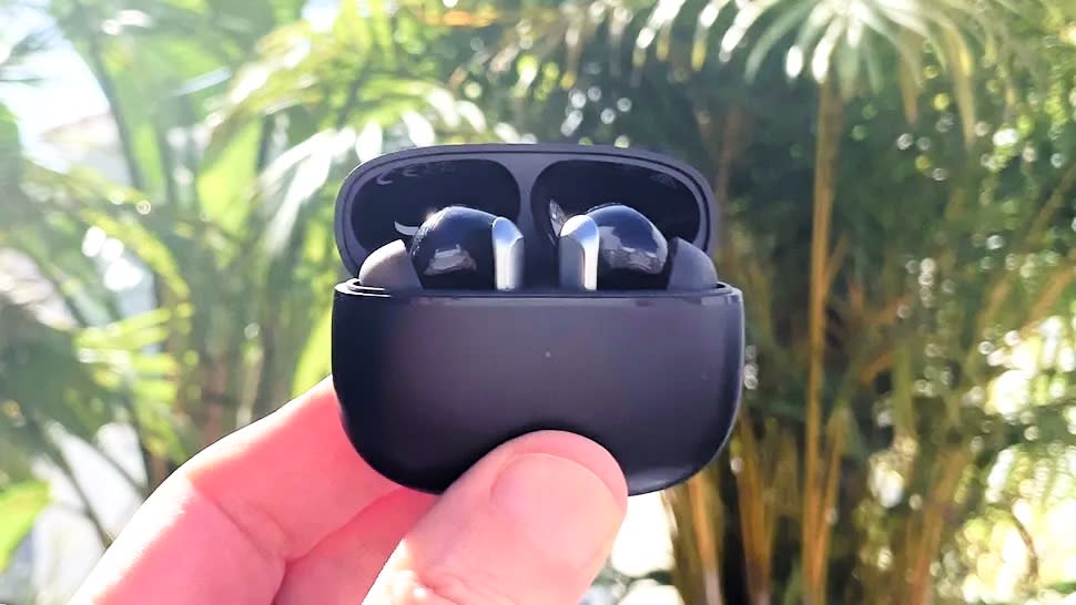  EarFun Air 2 in charging case held  between fingers in an outdoor setting with plants in the background. 