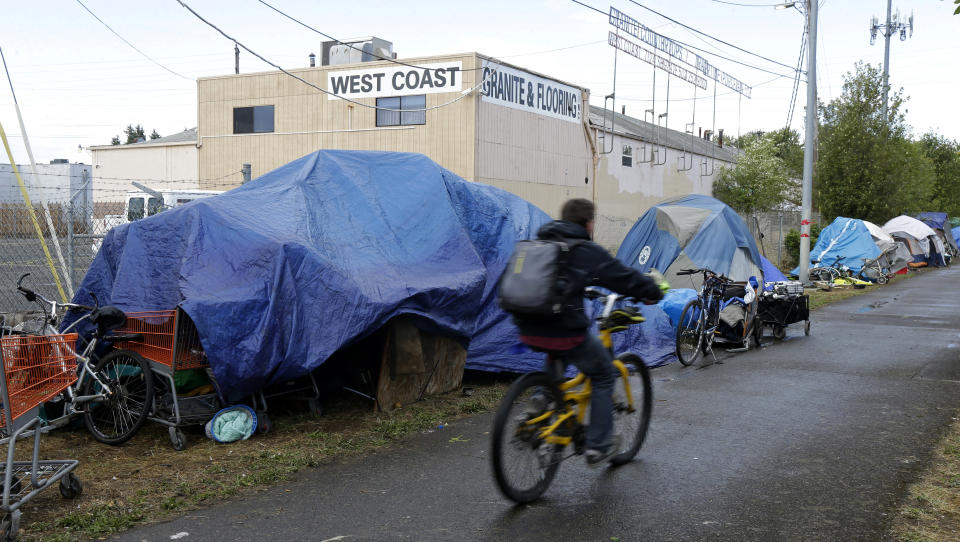 FILE - In this Sept. 19, 2017, photo, a person cycles past tents set up along a pathway in Portland, Ore. Voters in Portland will be asked Tuesday, May 19, 2020 to approve taxes on personal income and business profits that would raise $2.5 billion over a decade to fight homelessness even as Oregon grapples with the coronavirus pandemic and its worst recession in decades. (AP Photo/Ted S. Warren)