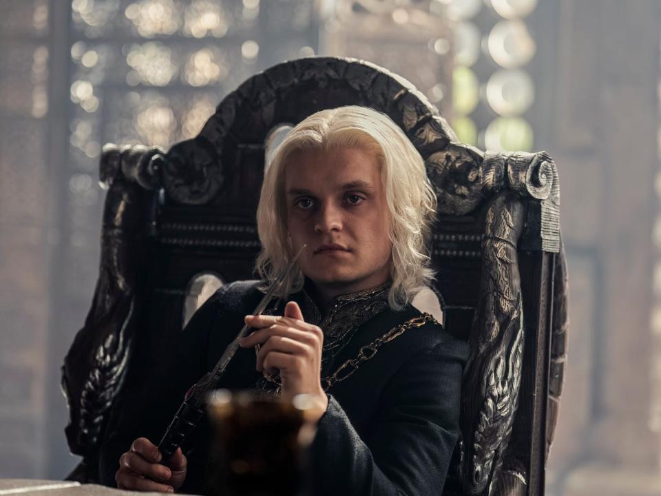 tom glynn-carney as aegon targaryen in house of the dragon. he's sitting in a high backed chair, playing with a steel dagger, and looking across a table