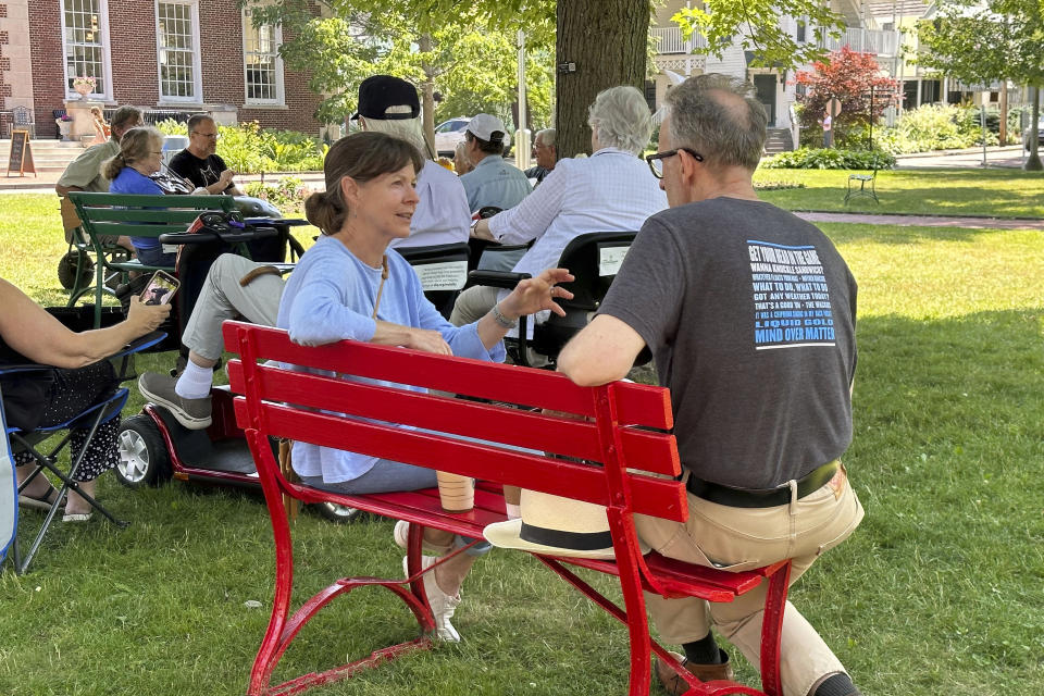 Mary Pat McFarland of Philadelphia, left, and Michael Crawford of Washington, D.C., talk while sitting on a red bench as a handful of musicians with violins, guitars and small harp, play an impromptu jam session beneath a tree nearby, on the grounds of the Chautauqua Institution, in Chautauqua, N.Y., Thursday, June 29, 2023. "It is a tragedy, it's heartbreaking," what happened to Salman Rushdie, Crawford said. "But I feel this is a very safe place and people are being patient with the increased security measures." (AP Photo/Carolyn Thompson)