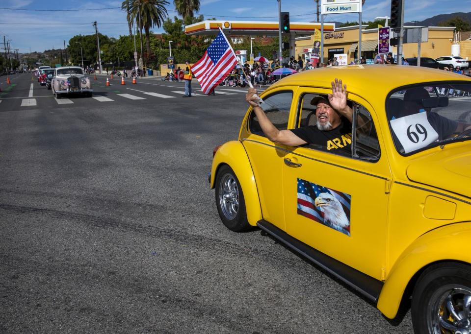 A man waves from a yellow VW bug with photo of a flag and eagle on the side door.
