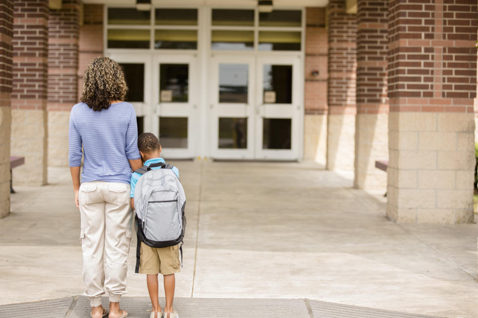 Seen from behind, an adult and child stand closely together a dozen yards from the entrance to what appears to be a school building. The child wears a backpack and rests their head on the adult's arm.