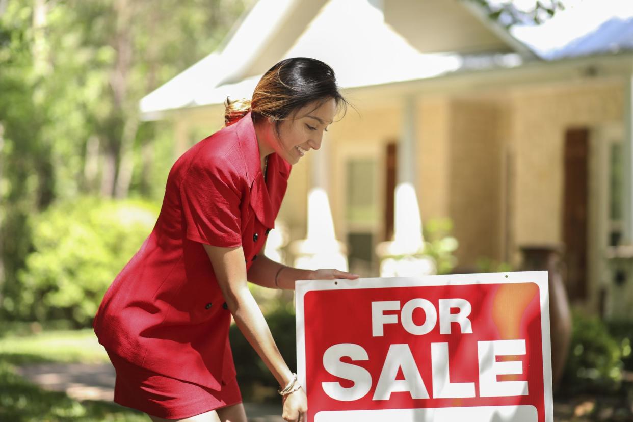 Female real estate agent putting a 'for sale' sign in the front yard of a home with lots of trees and shade in yard during spring with a blurred background of the yard