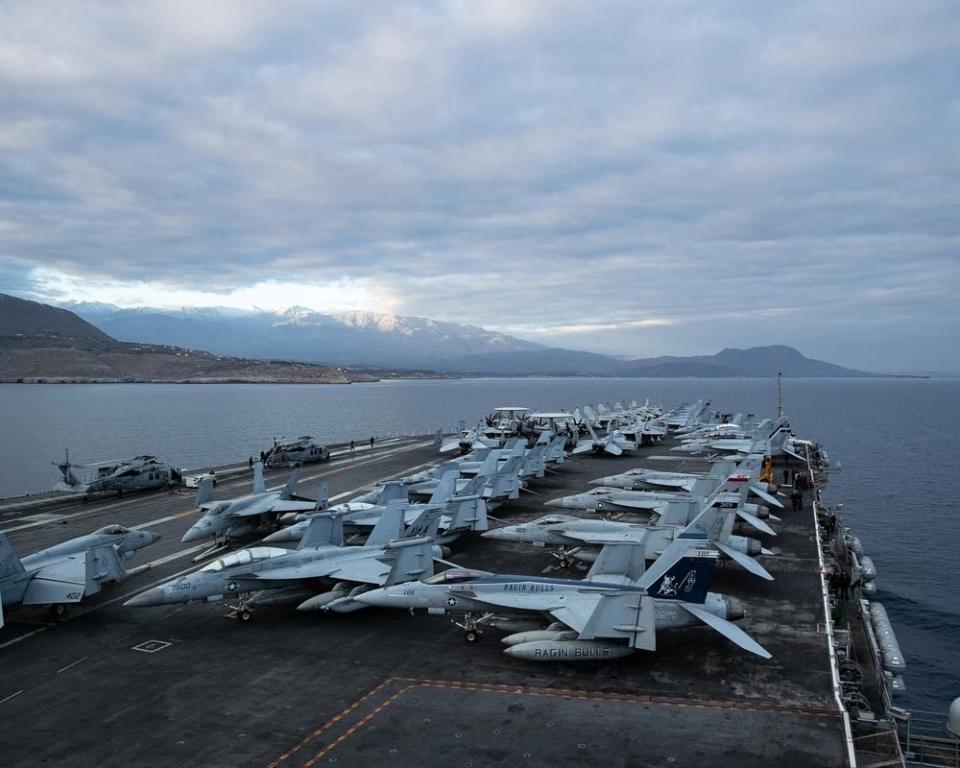 military aircraft are seen on the flight deck of a supercarrier with the coast of Greece in the background