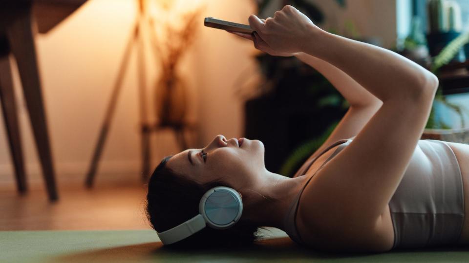 Woman laying down on a yoga mat holding phone above face, wearing headphones