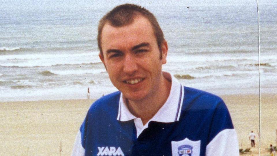 Martin has short brown hair and is wearing a blue and white Cardiff City shirt. Behind him is the beach and shoreline.