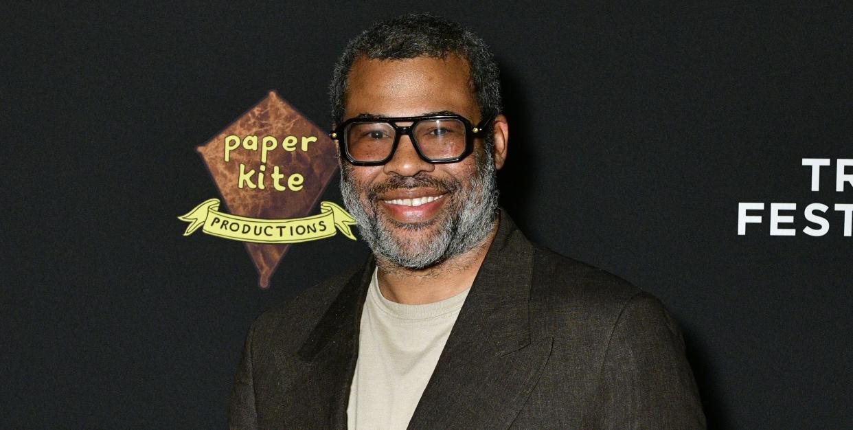 jordan peele, a man with black and grey hair and a black and grey beard, wearing black framed glasses, a tan shirt, and dark blazer while smiling for the camera