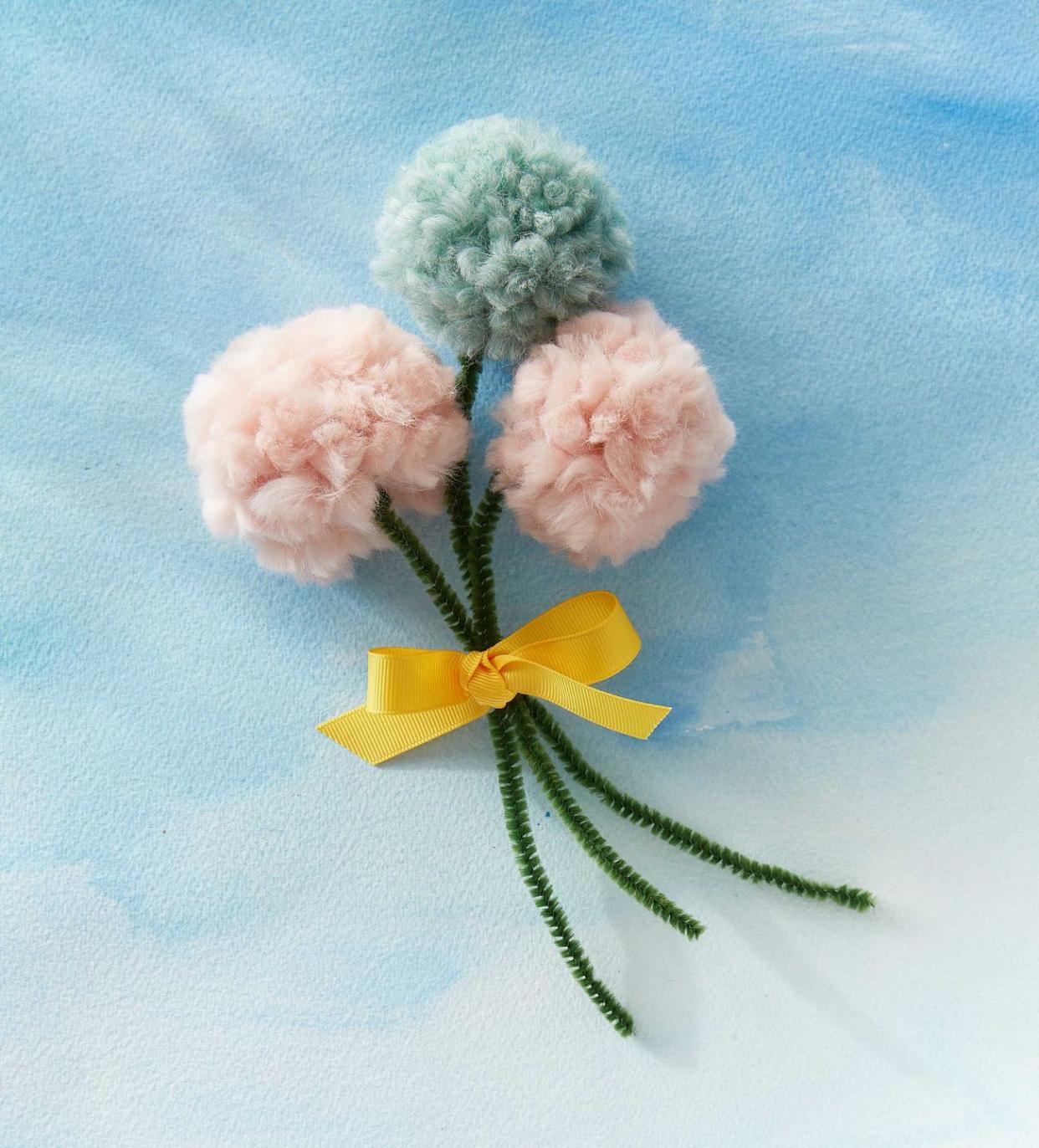 two pink and one pale blue yarn pom poms with green pipe cleaner stems tied into a mothers day bouquet with yellow ribbon