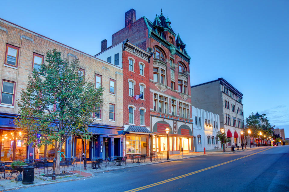 A quiet street with historic three-story buildings, including a brick building with a unique ornate roof and a few trees with outdoor seating areas along the sidewalk