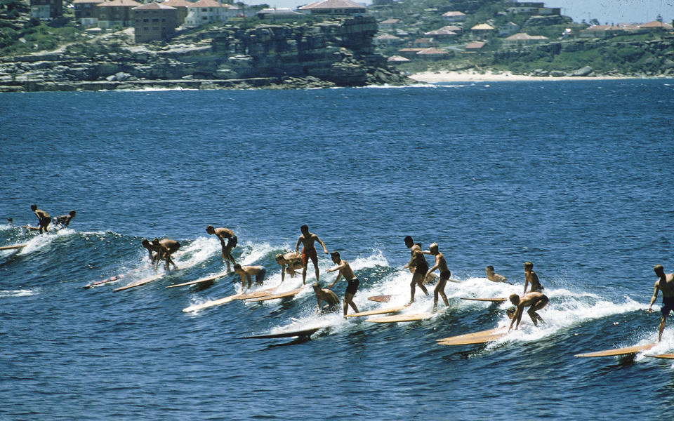 Off Manly Beach, near Sydney, Australia, a row of surfers ride a low wave back to shore.