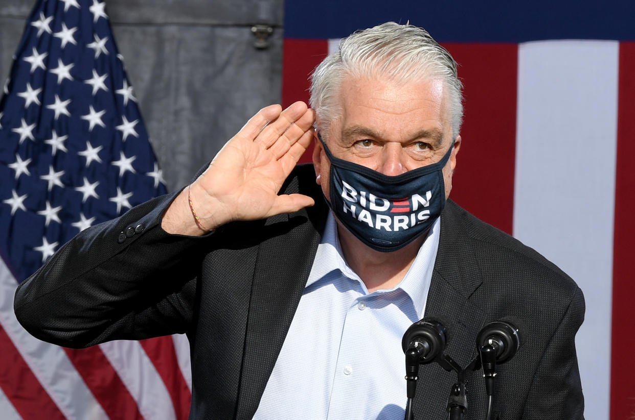 Nevada Gov. Steve Sisolak wears a Biden/Harris face mask and stands at a podium next to an American flag