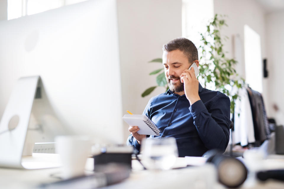 A man in a professional office setting, wearing a dress shirt, is sitting at a desk, holding a notepad, and speaking on a cellphone