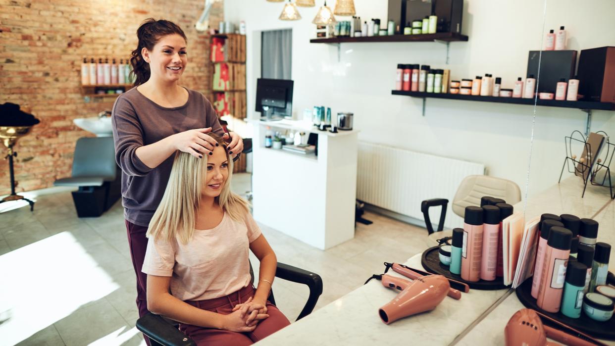 Young blonde woman smiling and looking at her reflection in a mirror while sitting in a salon chair during an appointment with her hairstylist.