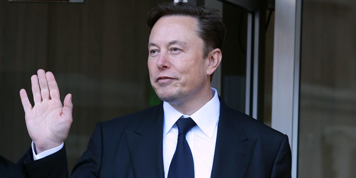 Elon Musk, wearing a black suit and tie, waves to photographers outside a courthouse