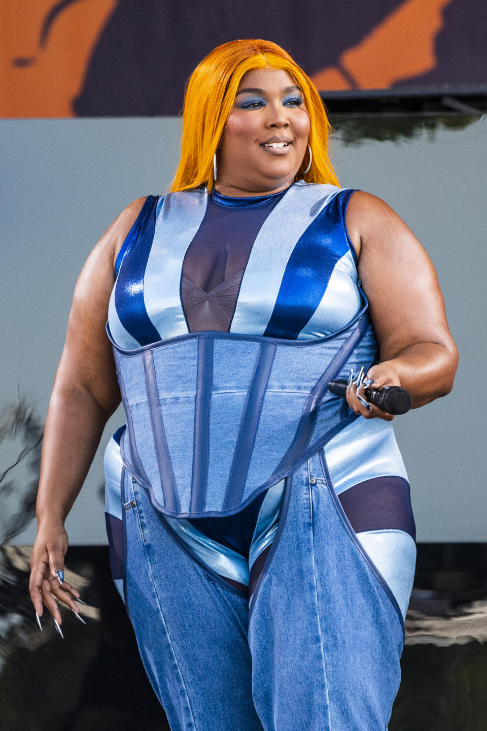 Lizzo performs on stage, wearing a blue jumpsuit with structured corset details and holding a microphone