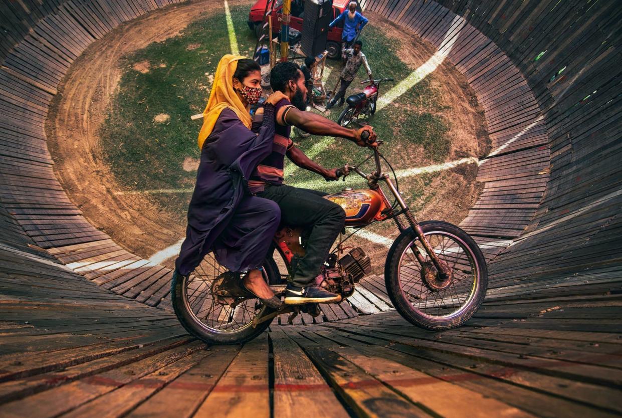 An aerial view looking into the "Well Of Death," where a man and woman ride a motorcycle around a barrel shaped cylinder, nearly vertical to the ground.