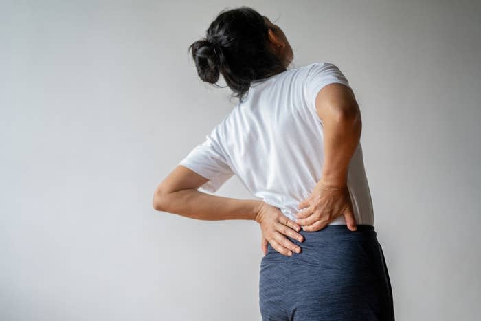 Person with long hair tied in a bun wearing a white shirt and dark pants holding their lower back, appearing to be in pain. No celebrities identified