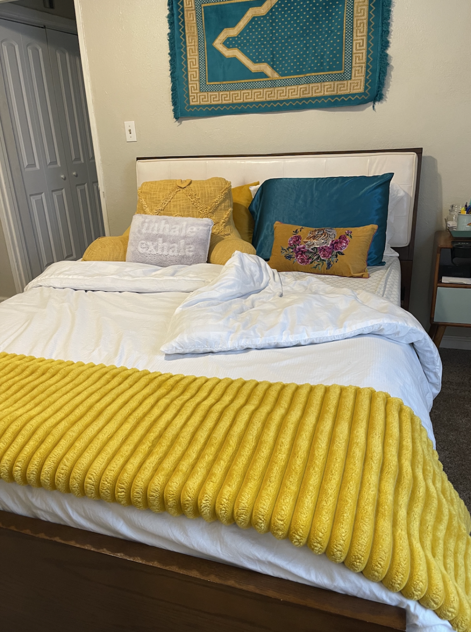 A neatly made bed with colorful pillows, a yellow throw blanket, and a decorative wall tapestry