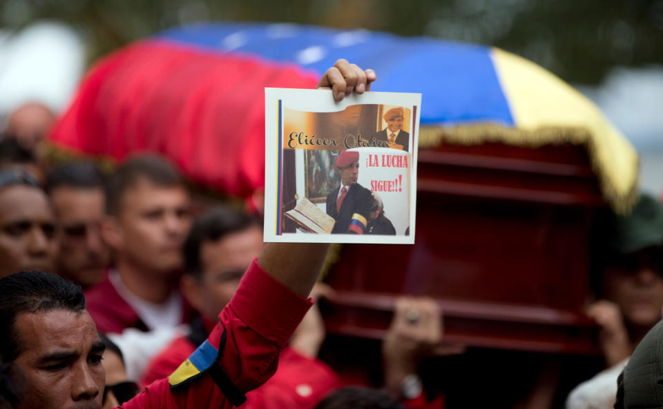 A person holds up a photograph of former Venezuelan Intelligence Chief Eliezer Otaiza as his flag-draped coffin is carried outside the National Assembly during a funeral procession in Caracas, Venezuela, Wednesday, April 30, 2014. Otaiza's body was dumped on the edge of Caracas Saturday after his vehicle was intercepted by a group of armed men. No motive has been established for the crime. As a young army officer, he backed Hugo Chavez's failed 1992 coup attempt and was responsible for his personal security when he was elected president in 1998. (AP Photo/Fernando Llano)