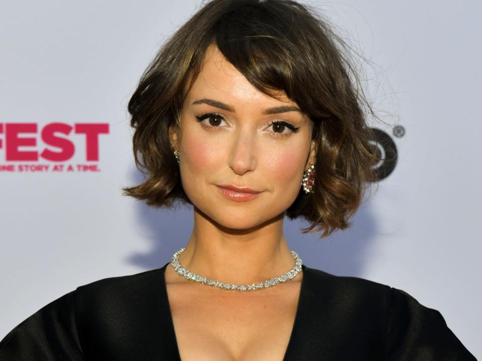 Actress Milana Vayntrub stands in front of a white background wearing a black dress and a sparkly necklace.