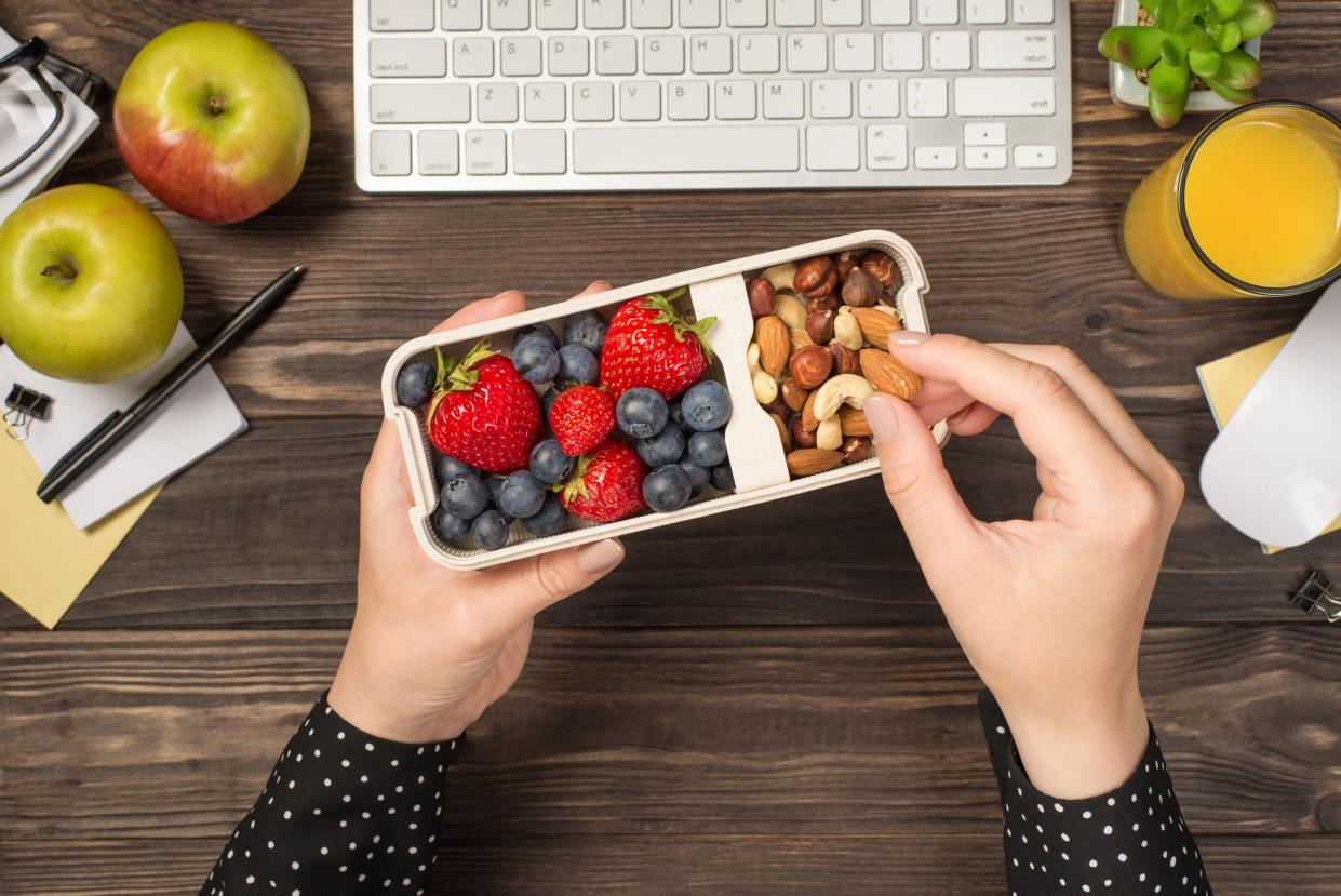 A close up top view of a person's hands picking up an almond out of a lunch box of berries and nuts, with an office desk in the background.