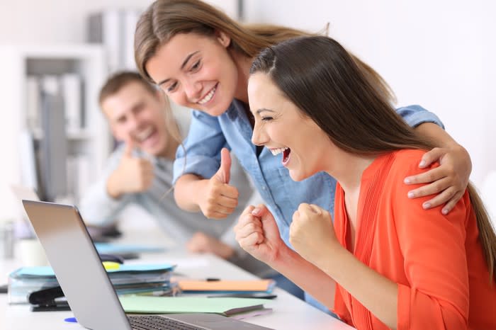 Two smiling office workers give thumbs-up encouragement to a colleague, who is cheering at her laptop.
