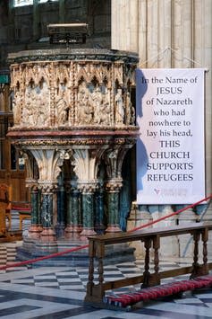 Ornate pulpit in the cathedral alongside which is a large banner hanging from a column in the name of Jesus of Nazareth proclaiming church support for refugees