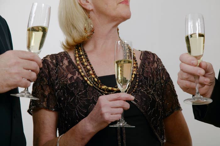 Three unidentified individuals toast with champagne flutes, featuring an older woman in a beaded necklace and lace attire in the center