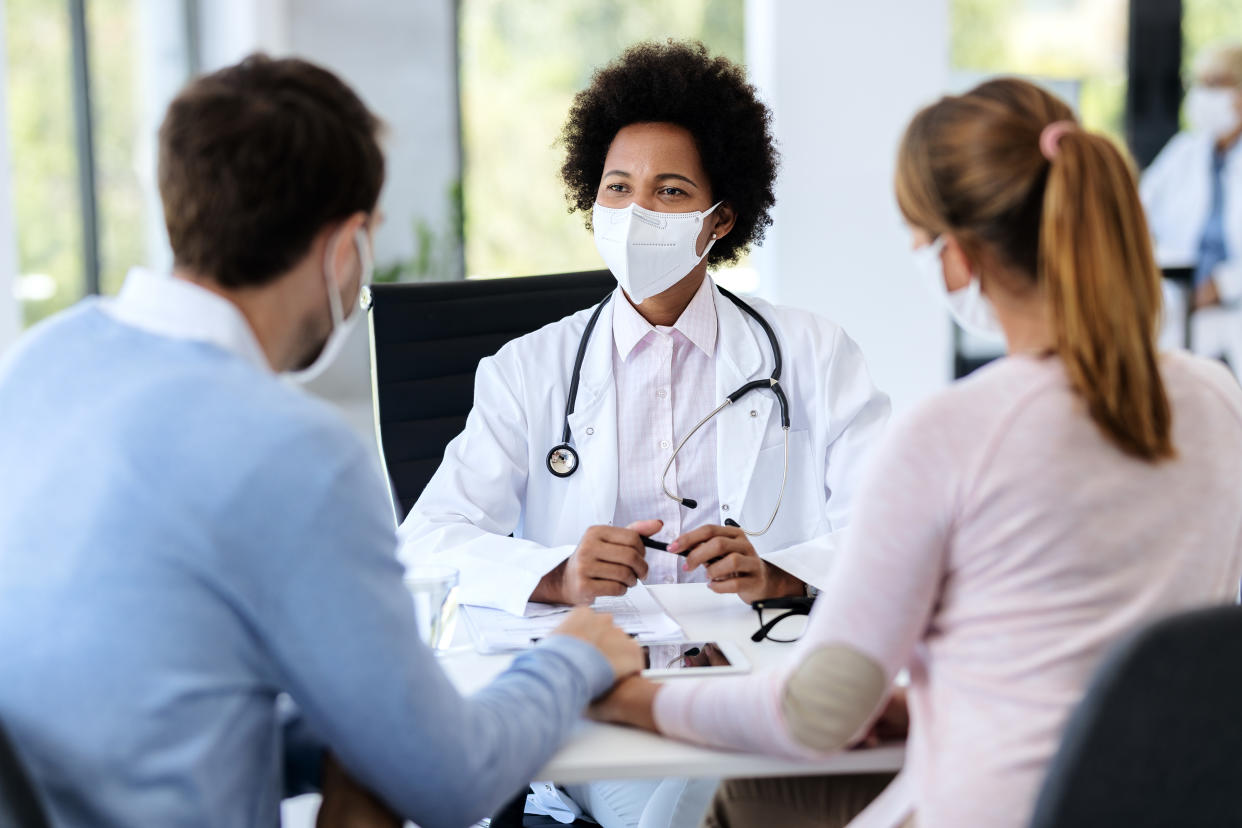 Black female doctor talking to a couple while wearing protective face mask during counseling at doctor's office.