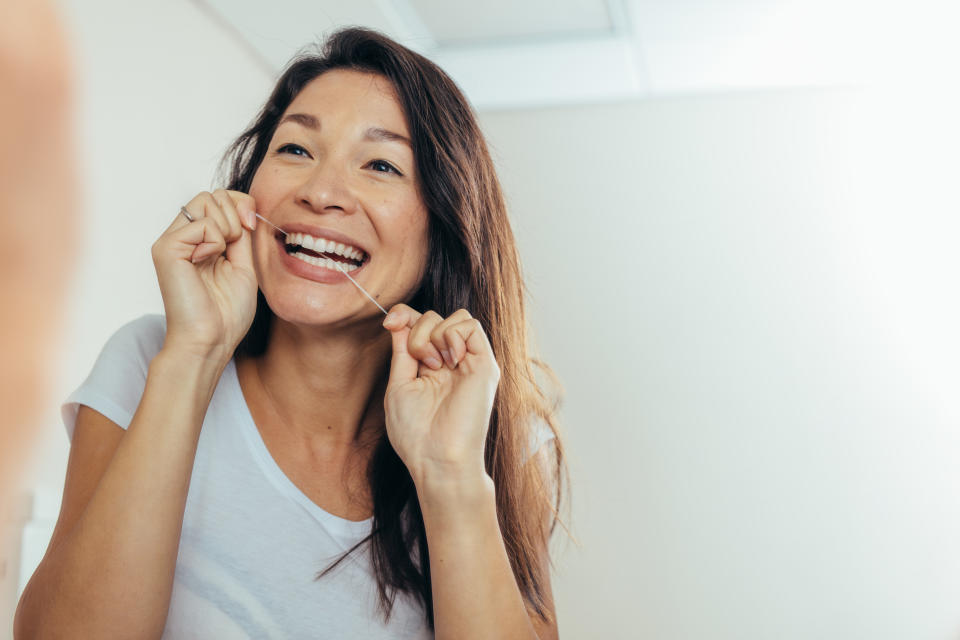 Woman looking in the bathroom mirror and using dental floss to clean her teeth. Reflection of woman in bathroom mirror while brushing teeth in morning.